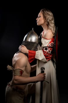 Young shirtless viking holding a sword staying on his knees and parting with his beautiful wife isolated profile shot on black background