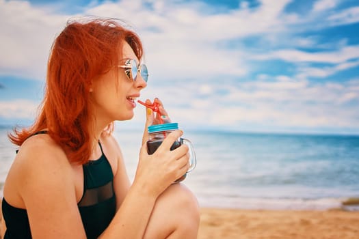 Calm lady in a swimsuit is sitting on beach with a cocktail and look away. Tropical alcohol beverage. Summer day with cloudy sky. Red-haired woman with sunglasses on vacation. Holidays at the sea.