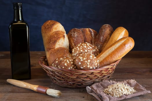 A basket of assorted baquetts and rolls with olive, a brush, and pile of oats.