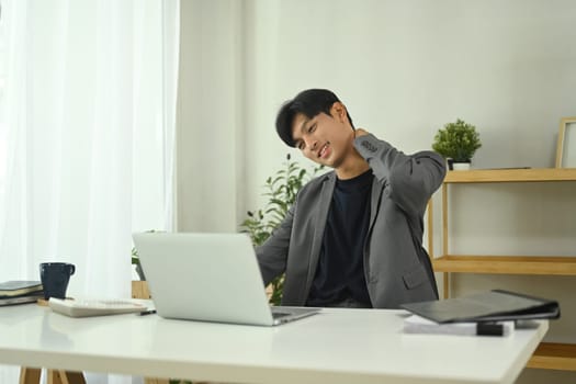 Young businessman in formalwear suffering from neck pain while working on a laptop at workstation.