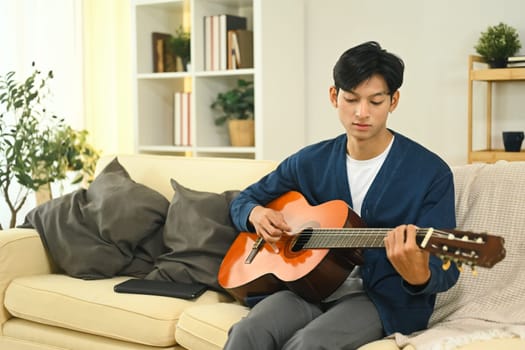 Young asian man relaxing and playing acoustic guitar in living room.