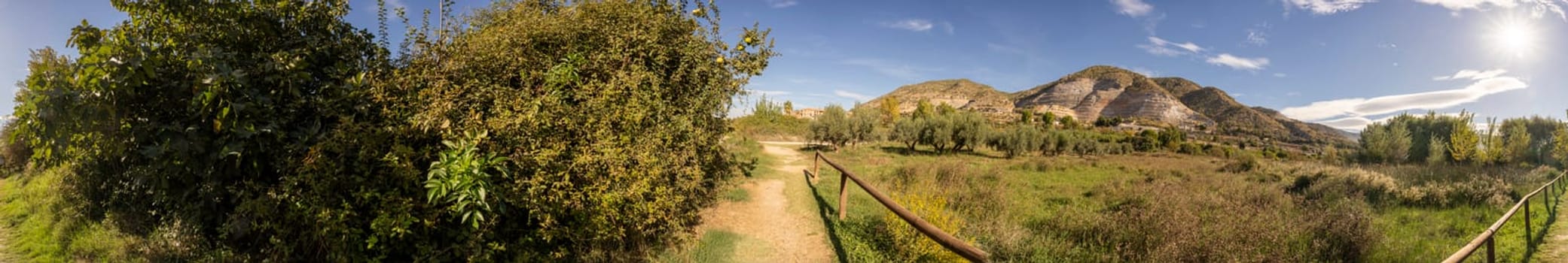 A stunning panoramic view showcases an abundance of dense greenery, along with a serene and tranquil pathway, framed by majestic mountains rising majestically beneath a clear, vast blue sky