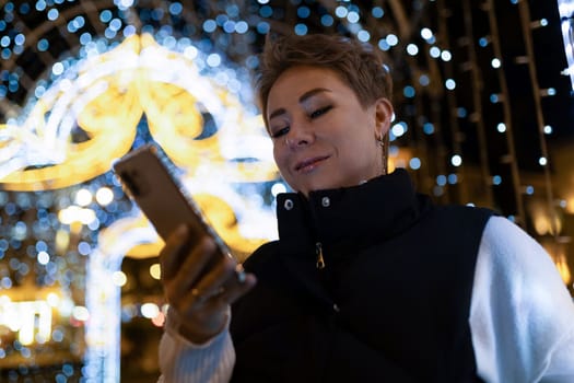 A woman with short brown hair walks through a city at night. She is looking at her phone and is smiling. There are string lights in the background, out of focus.