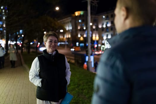 A married couple walks through a city at night. The woman is smiling and looking to the side. She is wearing a white shirt, a black vest, and a skirt.