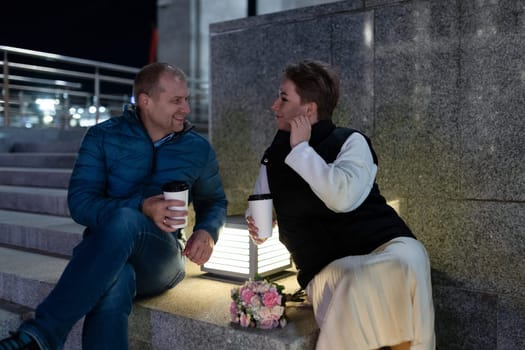 A man and a woman are sitting on steps in the city at night. The man is wearing a blue jacket and jeans, and he is holding a cup of coffee.