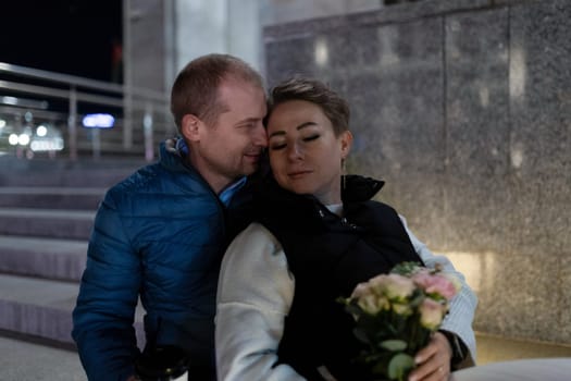 A man and woman walk together at night in the city. The man has his arm around the woman's waist, and she holds a bouquet of flowers.