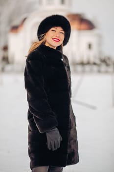 A beautiful woman in a black fur coat and a black hat has a lot of snow in winter, she is wearing red lipstick, smiling.