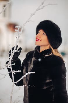 A beautiful woman in a black fur coat and a black hat has a lot of snow in winter, she is wearing red lipstick, smiling.