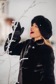 A beautiful woman in a black fur coat and a black hat has a lot of snow in winter, she is wearing red lipstick, smiling.