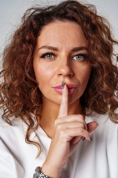A young woman with curly hair and blue eyes makes a silence gesture in a studio setting close up