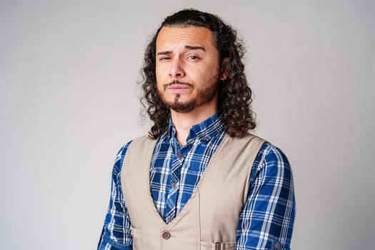 Confident man with curly hair wearing a plaid shirt and vest poses against a neutral background in a thoughtful expression