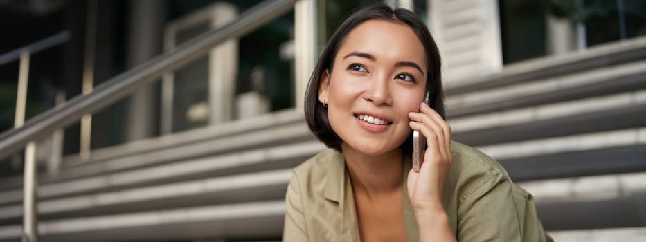 Portrait of asian girl smiles while talks on mobile phone. Young woman calling a friend, sitting on stairs. Technology concept