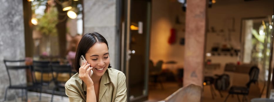 Portrait of young woman talking on mobile phone while typing, using laptop, working remotely from outdoo cafe, doing homework and calling.