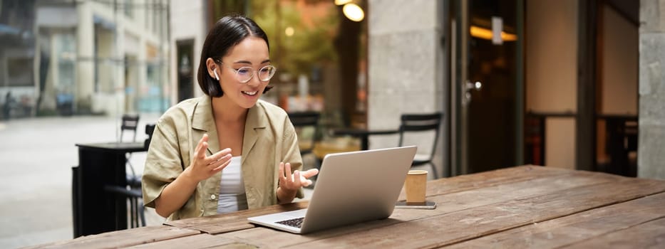 Young woman sitting on online meeting in outdoor cafe, talking to laptop camera, explaining something, drinking coffee. Digital nomad