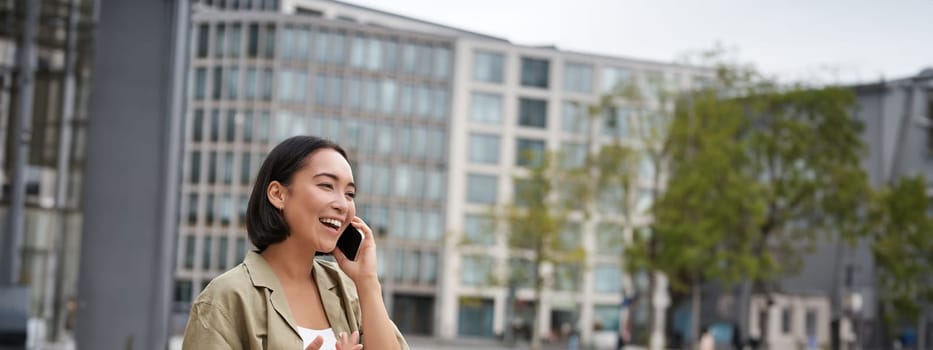 Vertical shot of smiling stylish asian girl, having a telephone conversation and walking on street. Young woman talking on mobile phone.