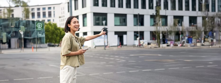 Vertical shot of young woman trying catch taxi, waving at driver on road, holding smartphone with car sharing app, standing on city street.
