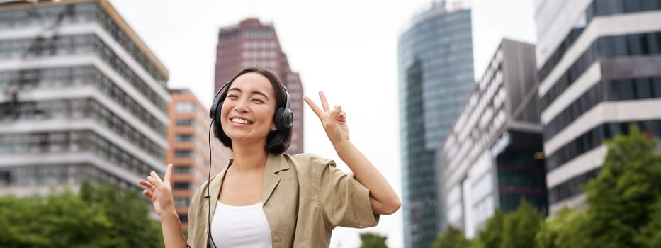 Happy asian woman in headphones, listening music and dancing on street of city centre, smiling with hands up.