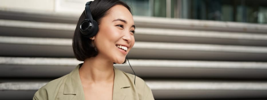 Smiling asian girl, laughing, listening music in headphones, sitting outdoors. Uni student enjoying free time.