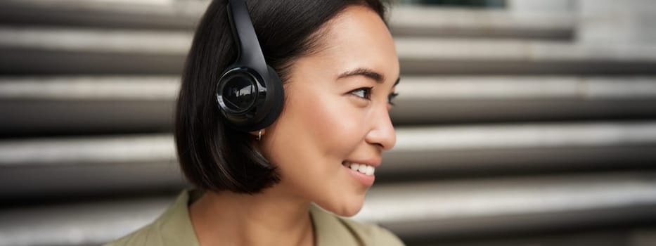 Close up portrait of smiling asian girl in headphones, listens to music outdoors, looking happy. People concept