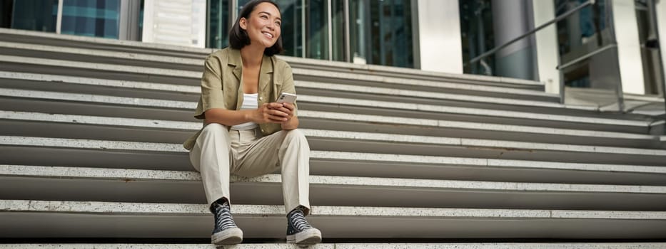 Portrait of happy smiling asian woman, sitting outdoors near building, using smartphone. Technology concept.