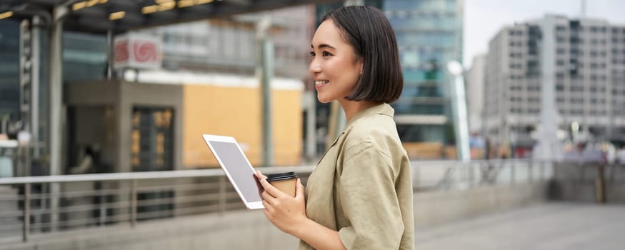 Modern people. Young asian woman with tablet, drinks coffee on street, using app, smiling.