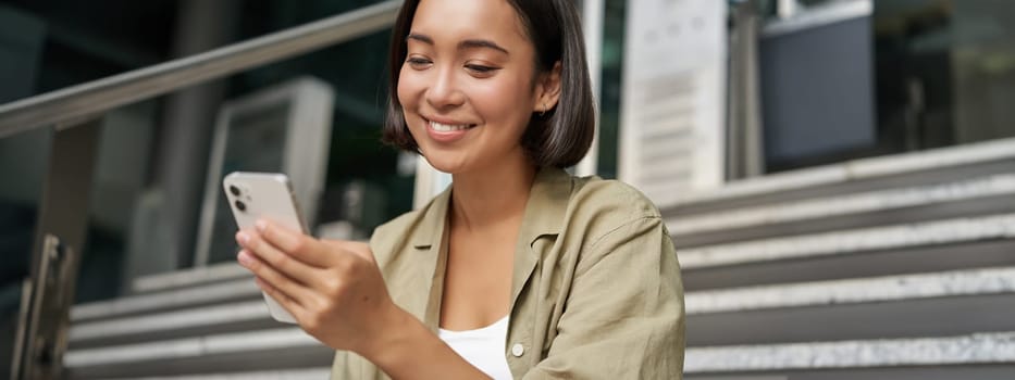 Portrait of asian girl takes selfie on mobile phone. Korean woman smiling, video chat on smartphone app while sits outside on stairs.