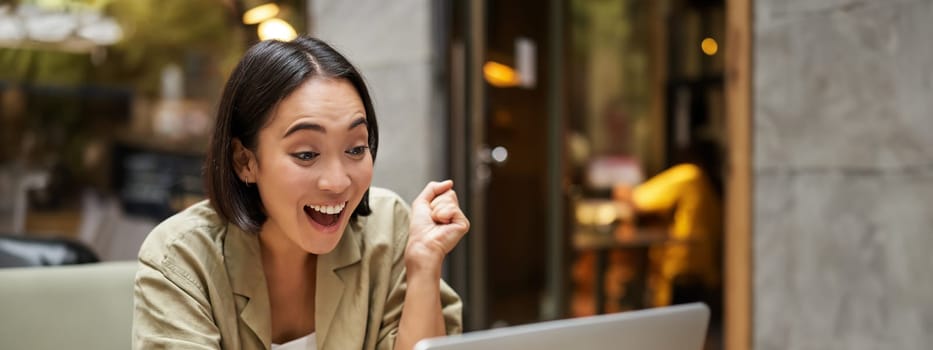 Vertical shot of happy girl talking on video call, looks at laptop, having online meeting, sitting in outdoor cafe.