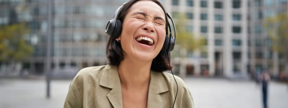 Portrait of happy asian woman in headphones, enjoys music while walking in city, smiling and laughing.