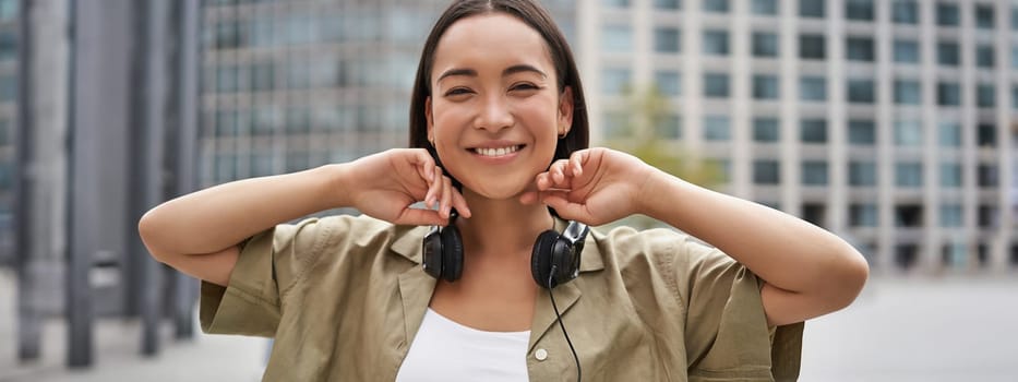 Portrait of smiling asian girl with headphones, posing in city centre, listening music.