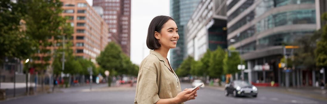 Young asian woman exploring city with smartphone app, holding mobile phone and walking on screet.