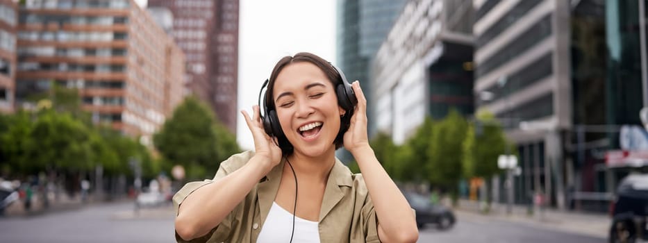 Dancing girl feeling happy in city. Asian woman dancing and listening music in headphones, posing on street.