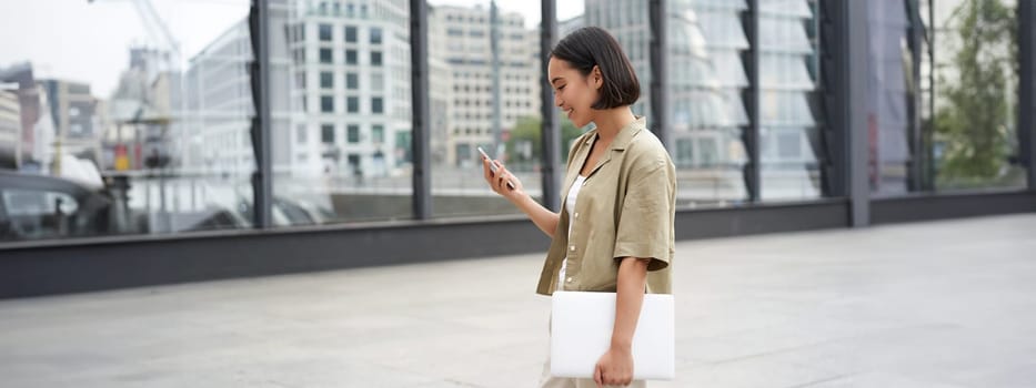 Young asian woman with laptop, walking on street and texting message, smiling while looking at smartphone.