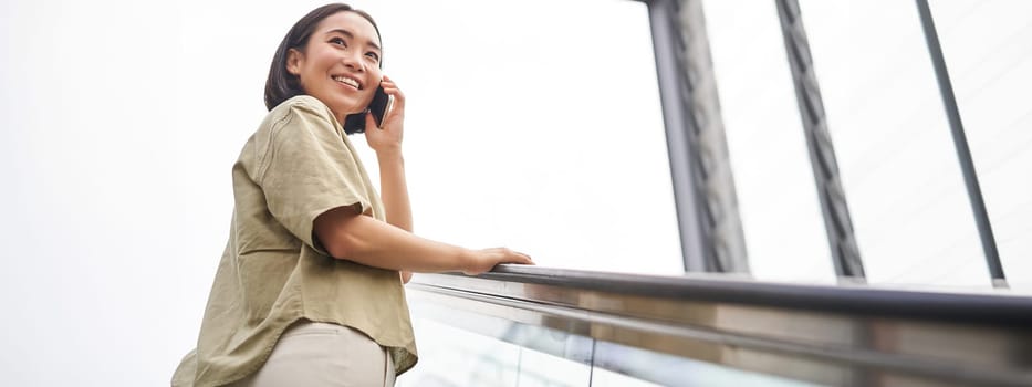 Happy asian woman on escalator, talking on mobile phone, meeting someone in city. Copy space