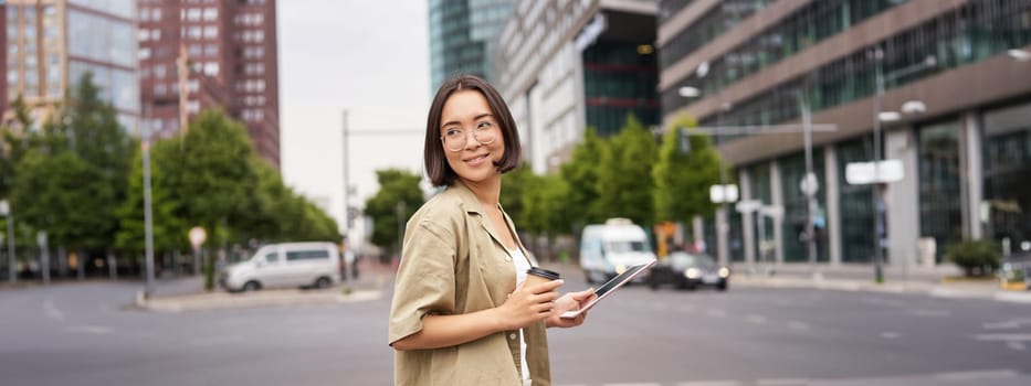 Happy woman exploring the city. Young korean girl holds tablet, drinks coffee and walks along street with big smile on her face.