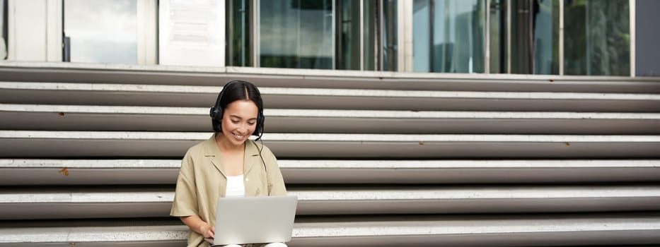 Smiling asian girl in headphones, works on laptop, digital nomad using computer on remote, sitting on stairs outdoors.