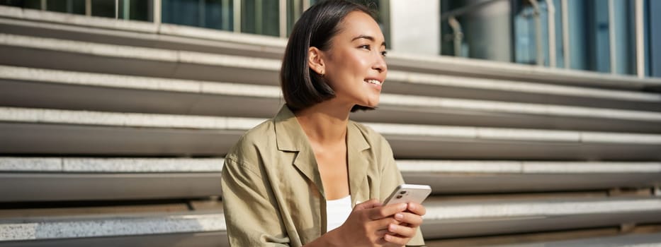 People and technology. Smiling beautiful asian woman sitting on stairs in city, holding mobile phone. Girl with smartphone rests outside.