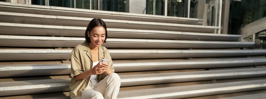 Beautiful asian girl sitting on stairs outside building, using mobile phone, looking at smartphone app and smiling.