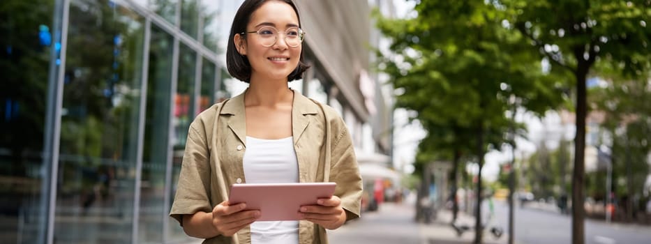 Portrait of young female student, standing near building on street, holding tablet and smiling.