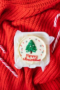 A small cake with the words Merry Christmas and a drawing of a Christmas tree lies on a red blanket along with candy canes. View from above