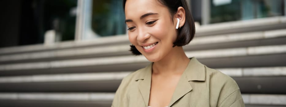 Portrait of smiling asian girl listens music, podast in wireless earphones, using headphones outdoors, sitting on street.