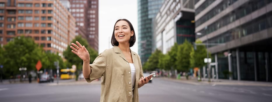 Happy asian girl passing by friend and waving at them on street, saying hello while walking in city, holding smartphone.