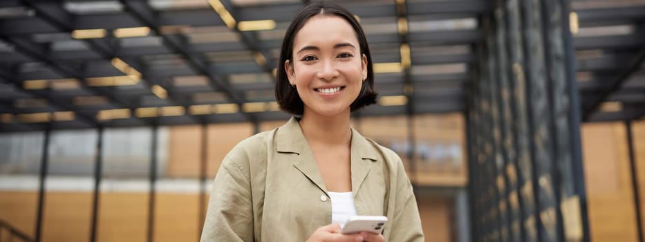 Technology and people. Smiling asian girl with mobile phone, using telephone and walking in city in daylight.