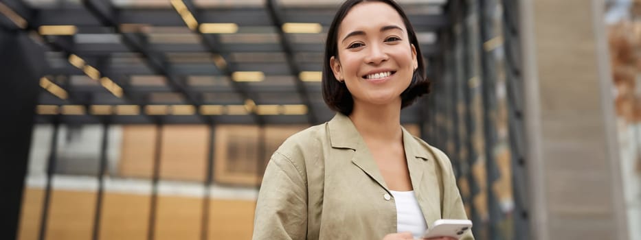 Technology and people. Smiling asian girl with mobile phone, using telephone and walking in city in daylight.