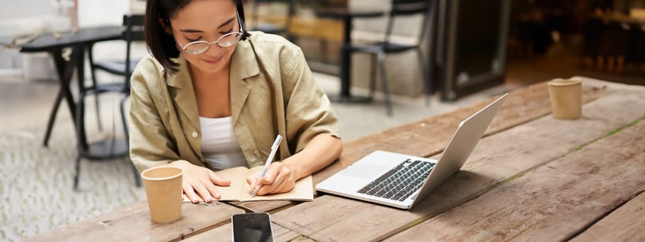 Portrait of concentrated girl doing homework, attending online course, making notes while working on laptop from cafe.