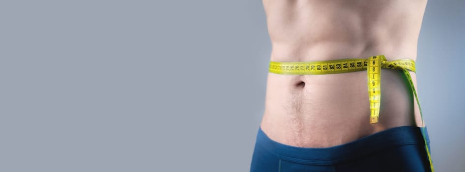 Male torso and measuring tape on a gray background. Unrecognizable muscular man in shorts taking measurements of his body.