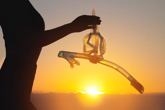 beach adventure, hand silhouette with snorkeling equipment. unrecognizable woman holding underwater mask and snorkel in her hand at sunset.