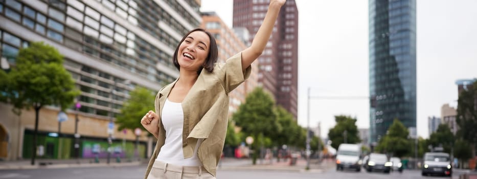Portrait of happy asian woman, dancing and feeling joy, triumphing, raising hand up in victory gesture, celebrating on streets.