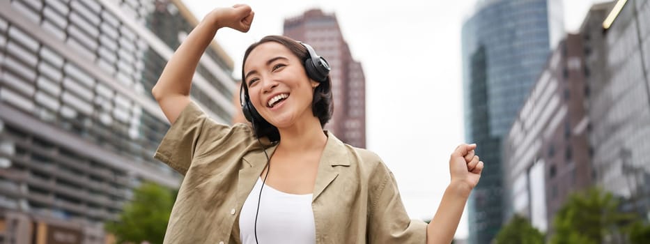 Dancing asian girl listening music in headphones, feeling happy in city, enjoying outdoors.