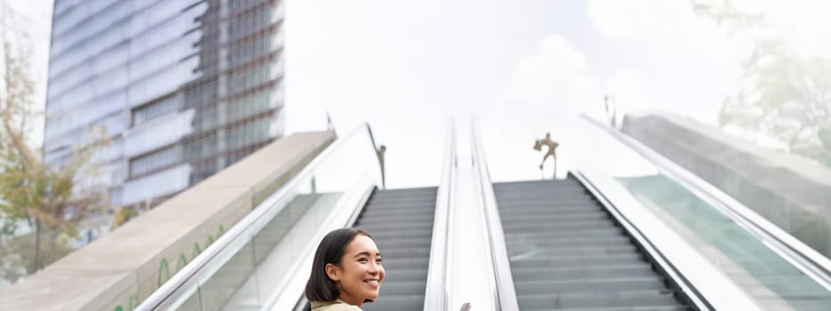 Young asian girl going up on an escalator, holding smartphone, smiling while walking in city. Copy space