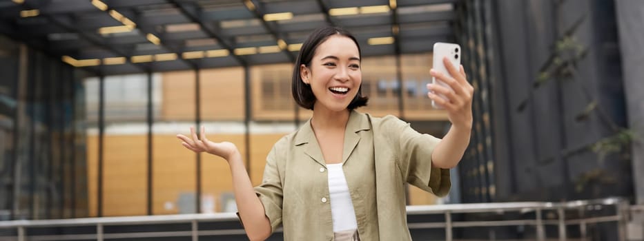 Happy asian girl shows something behind her during video call, demonstrating smth, standing on street.